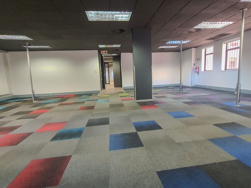 Open-plan commercial office space featuring a suspended ceiling, recessed lighting, structural columns, and multi-colored carpet tiles.
