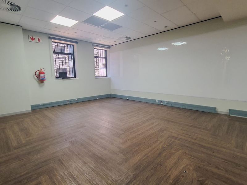 A clean, vacant office space featuring herringbone vinyl flooring, a large wall-mounted whiteboard, recessed ceiling lighting, and security bars on the windows.