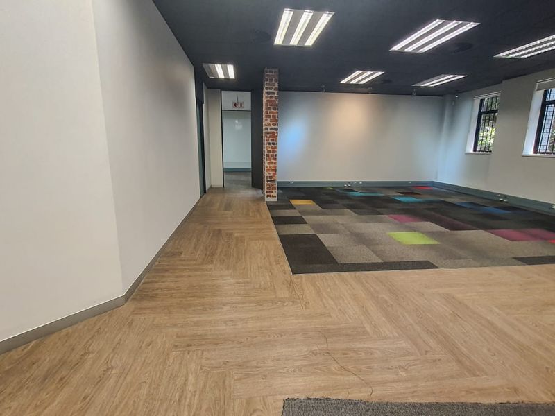 Spacious open-plan office area featuring a mix of herringbone vinyl and patterned carpet tile flooring, exposed brick column, and recessed ceiling lighting.