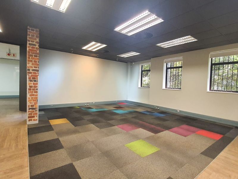 A spacious, empty commercial office area featuring a decorative exposed brick pillar, modern carpet tiles, and recessed lighting with secure window bars.