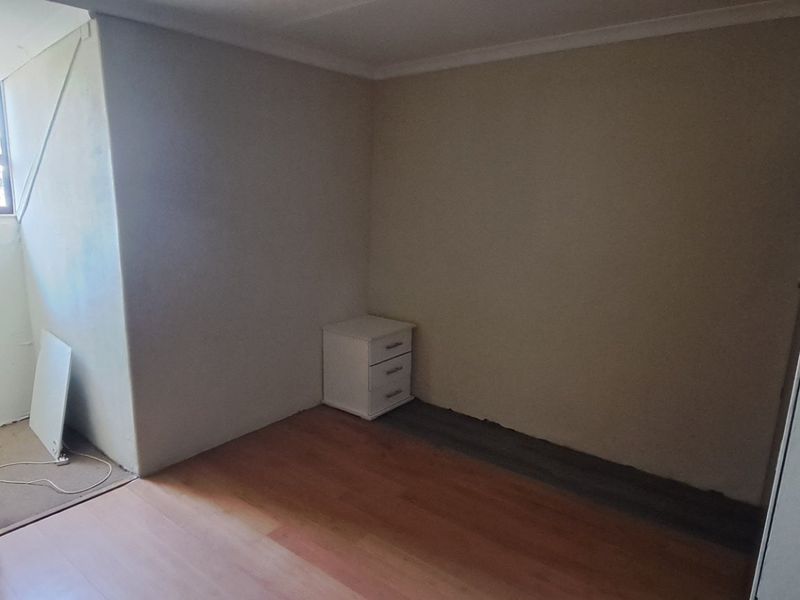 A sparsely furnished bedroom featuring laminate flooring and a casement window, requiring minor finishing touches to the flooring and wall surfaces.