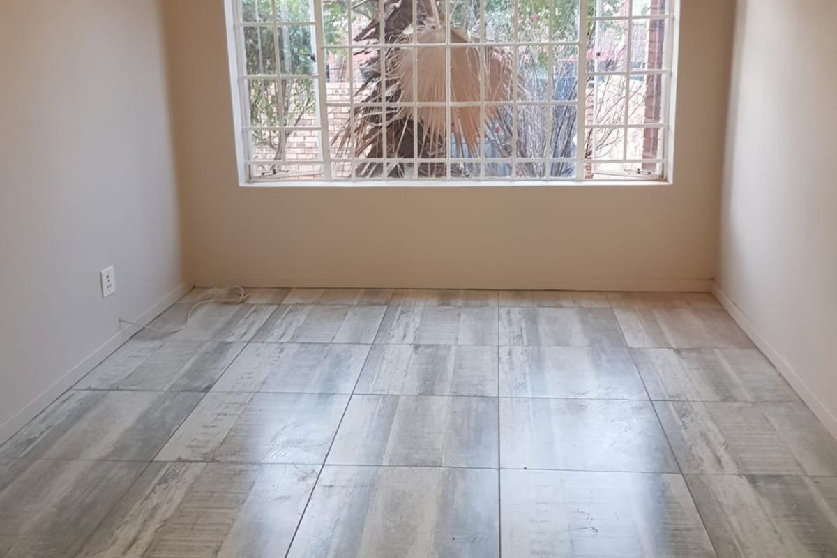 A bright, empty bedroom featuring modern wood-look tiled flooring and a large window fitted with security bars.