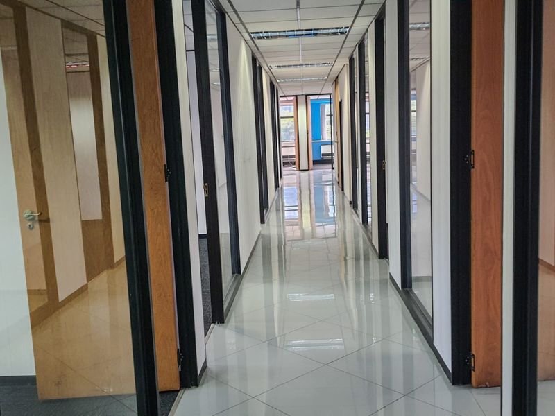 A bright, modern commercial office hallway featuring polished tiled flooring, glass partition walls, and a suspended ceiling with recessed lighting.