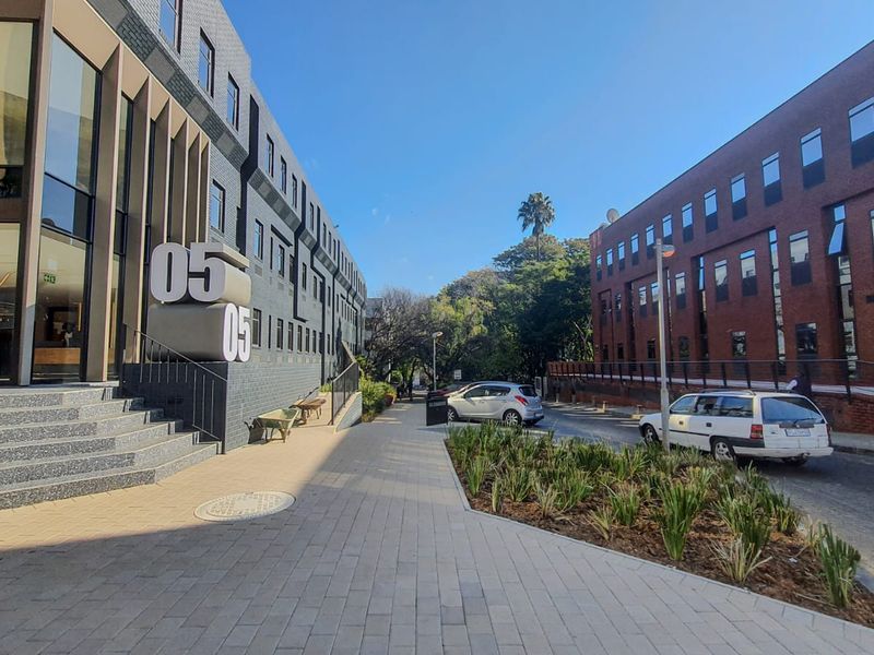 Modern commercial office building exterior featuring a paved pedestrian walkway, landscaped garden beds, and a prominent glass entrance facade.