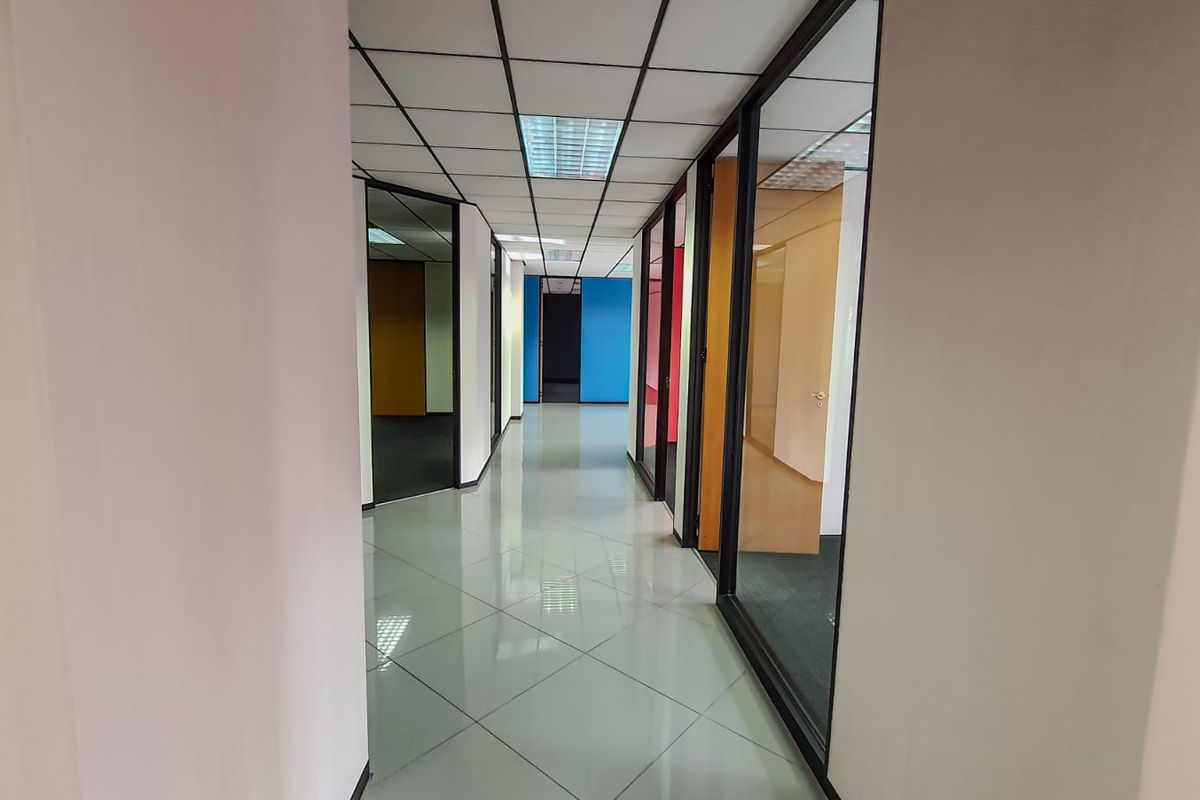 A bright commercial hallway featuring tiled flooring, suspended ceilings with recessed lighting, and glass partition walls leading to multiple office spaces.