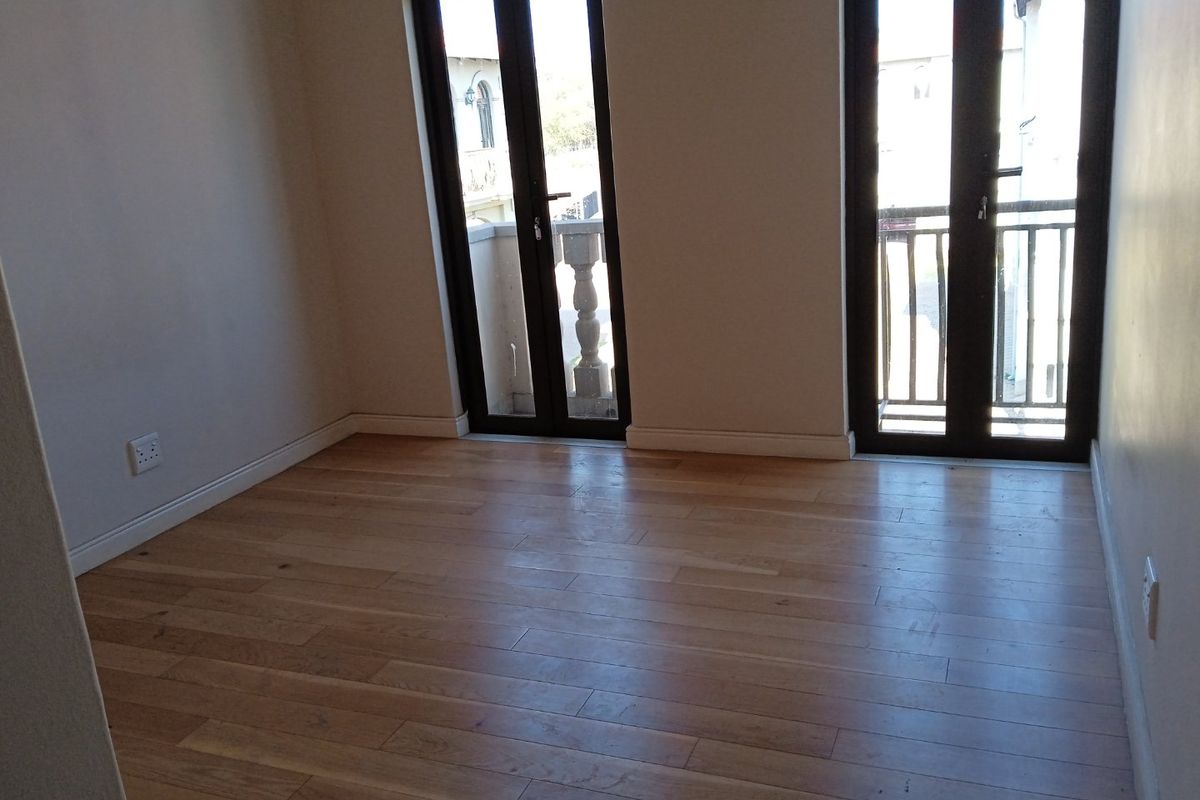 A bright, empty bedroom featuring polished hardwood flooring and double french doors leading to a balcony.