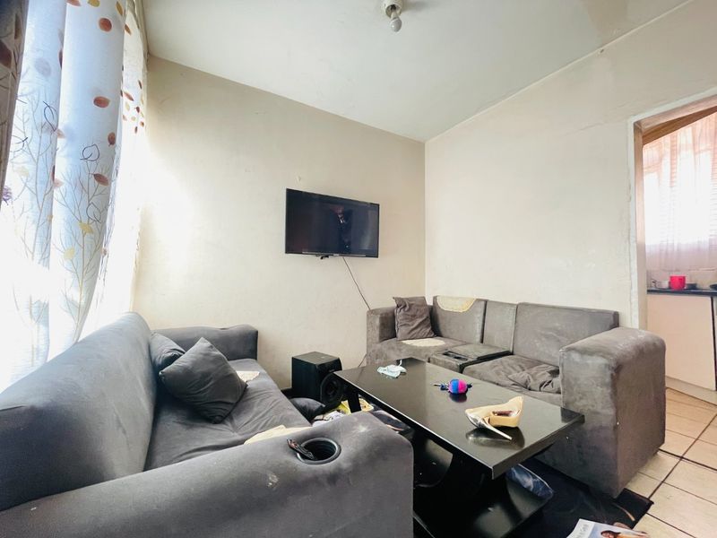 A compact living room featuring tiled flooring and wall-mounted television space, currently showing signs of wear on the walls and ceiling.