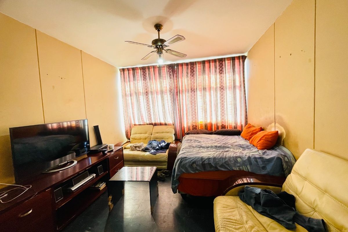 A bedroom featuring a ceiling fan and large window with curtains, currently used as a multi-purpose living and sleeping space.