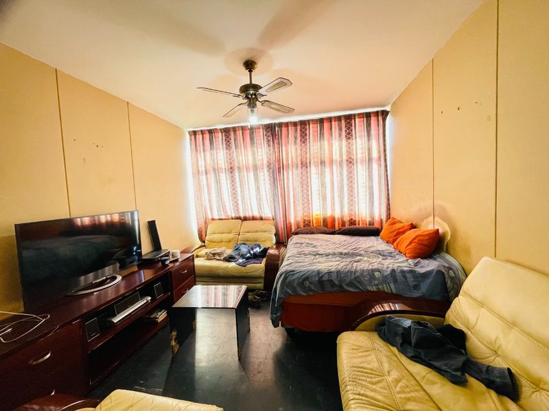 A bedroom featuring a ceiling fan and large window with curtains, currently used as a multi-purpose living and sleeping space.
