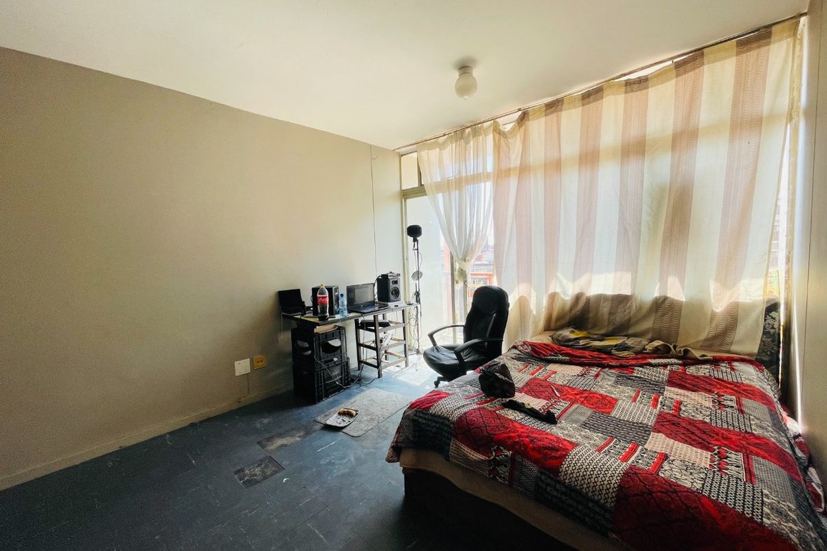 A bedroom featuring large floor-to-ceiling windows with access to a balcony, providing natural light. The room requires cosmetic updates to flooring and wall finishes.
