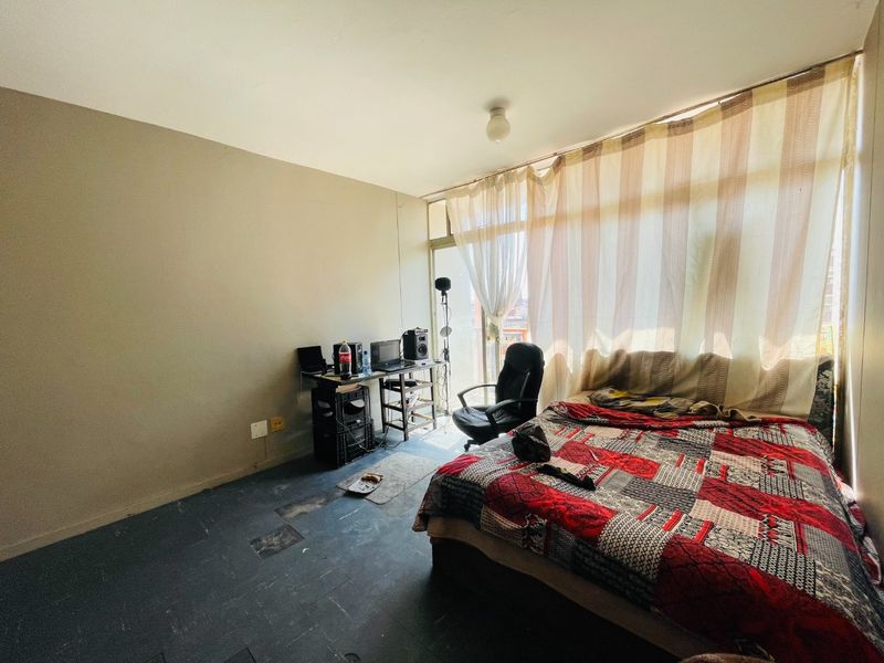 A bedroom featuring large floor-to-ceiling windows with access to a balcony, providing natural light. The room requires cosmetic updates to flooring and wall finishes.