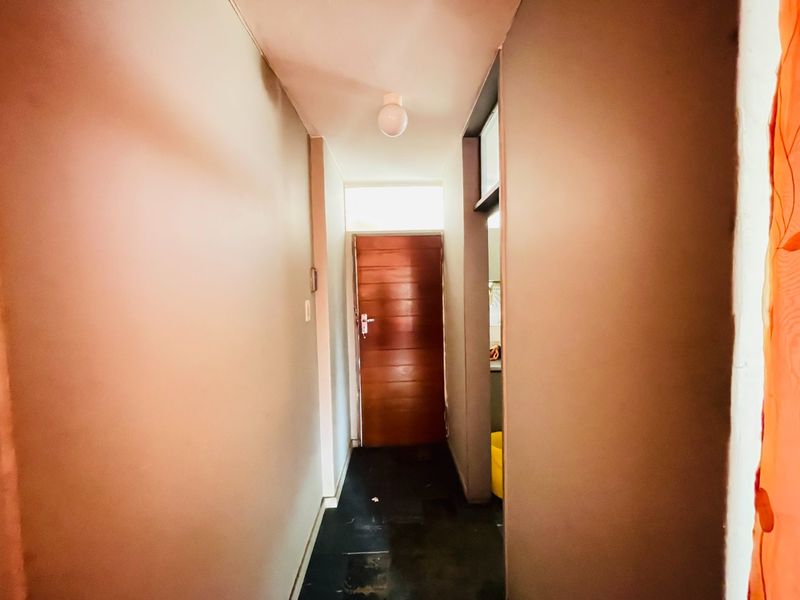 A narrow interior hallway featuring a wooden entrance door, overhead lighting, and a transom window, showing signs of wear on the flooring and walls.