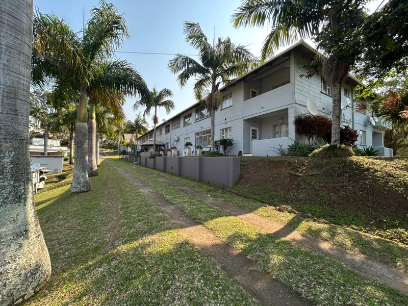 Exterior view of a multi-story apartment complex featuring balconies, a concrete retaining wall, and established palm trees in a landscaped garden setting.