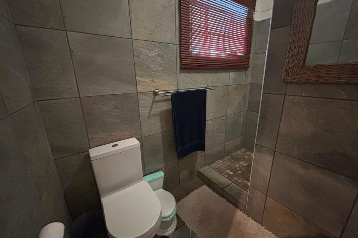 A compact bathroom featuring floor-to-ceiling stone-look tiling, a walk-in shower area, and a standard toilet with a window for natural light.