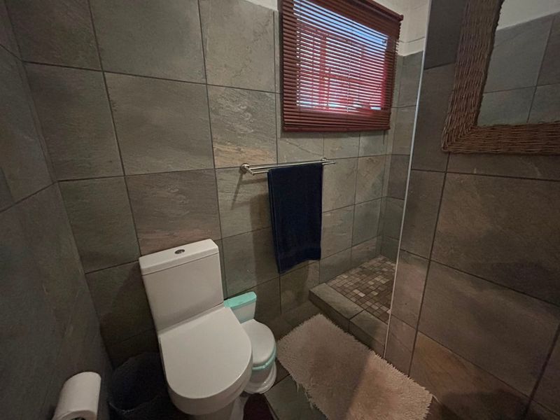 A compact bathroom featuring floor-to-ceiling stone-look tiling, a walk-in shower area, and a standard toilet with a window for natural light.
