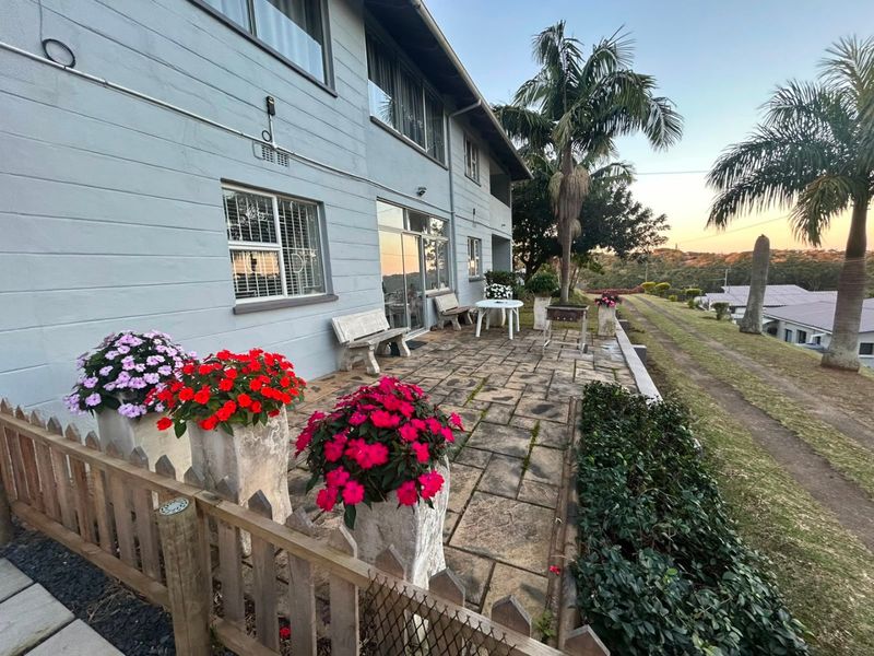 A paved outdoor patio area featuring a timber picket fence, sliding glass door access, and exterior wall lighting, overlooking the surrounding property grounds.