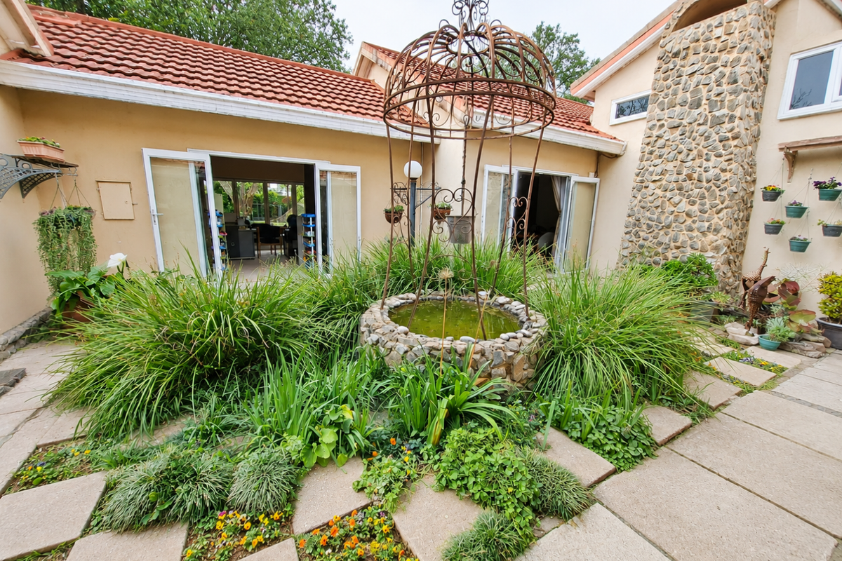 A rear courtyard featuring a stone-clad chimney, terracotta roof tiles, and a central stone water feature surrounded by paved patio areas and garden beds.