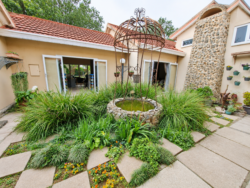 A rear courtyard featuring a stone-clad chimney, terracotta roof tiles, and a central stone water feature surrounded by paved patio areas and garden beds.