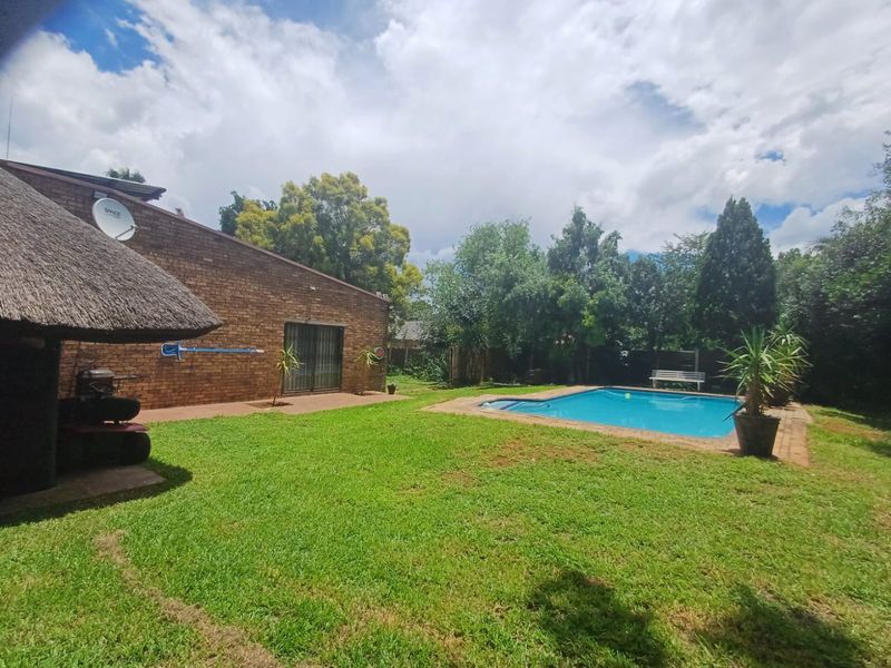 A backyard area featuring a swimming pool, a brick-walled house with a sliding door, and a thatched outdoor structure set within a lawned garden.