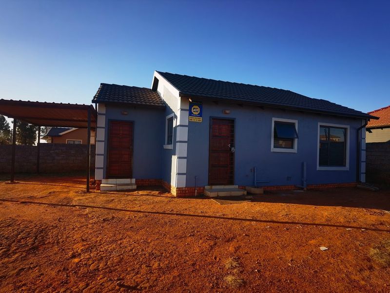 Front elevation of a single-story house featuring a tiled roof, attached carport, and security gates, set on an unpaved plot.