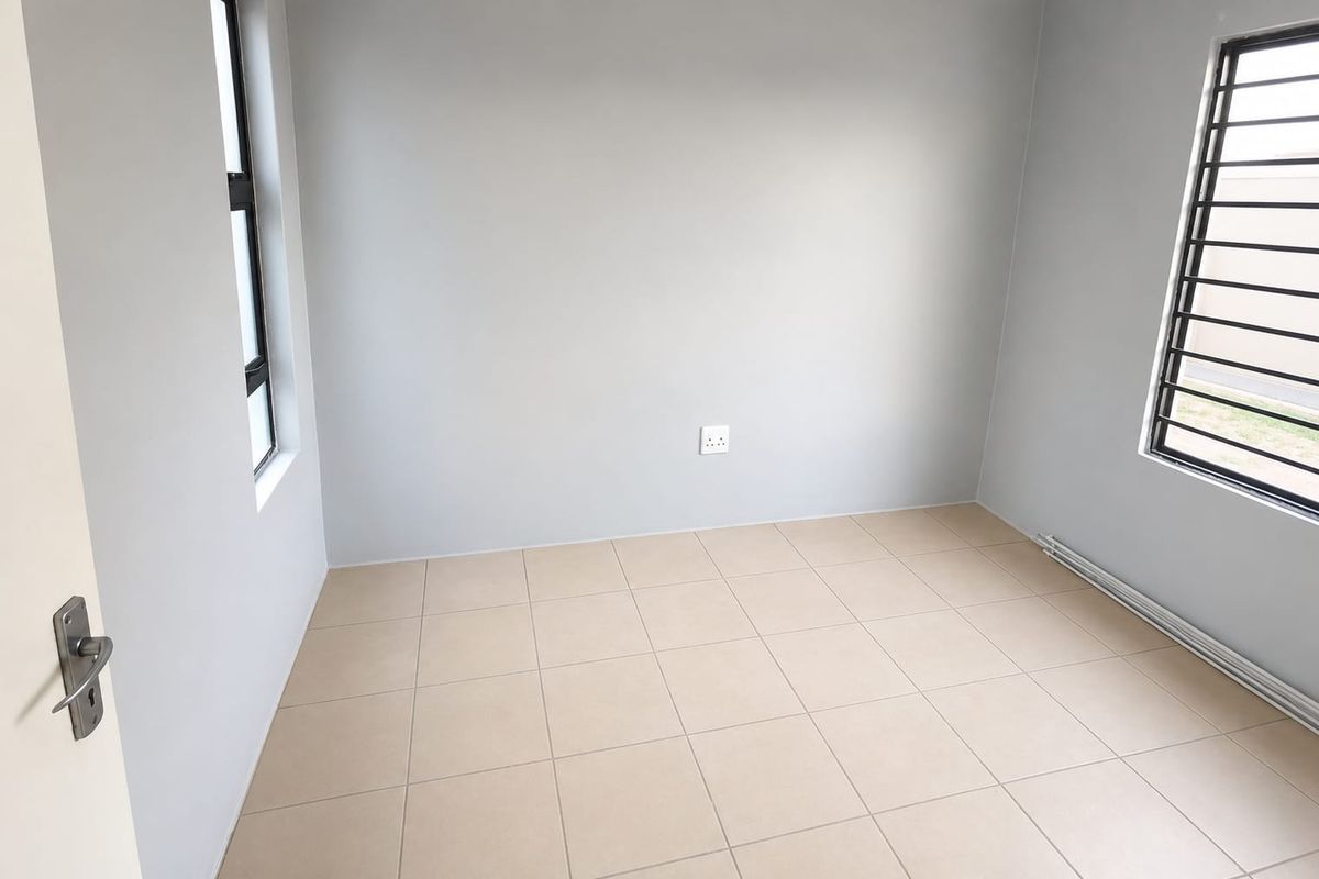 A clean, empty bedroom featuring neutral tiled flooring, light grey walls, and windows fitted with security bars.