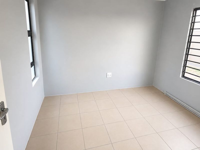 A clean, empty bedroom featuring neutral tiled flooring, light grey walls, and windows fitted with security bars.