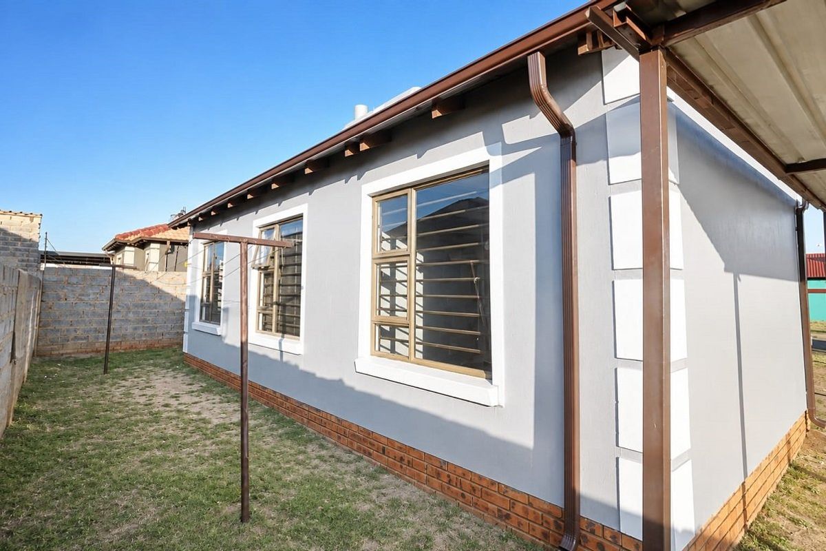 Rear exterior view of the property featuring a clean grey facade, brick base, aluminium windows with security bars, and a covered patio area.