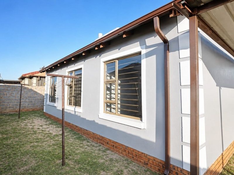 Rear exterior view of the property featuring a clean grey facade, brick base, aluminium windows with security bars, and a covered patio area.