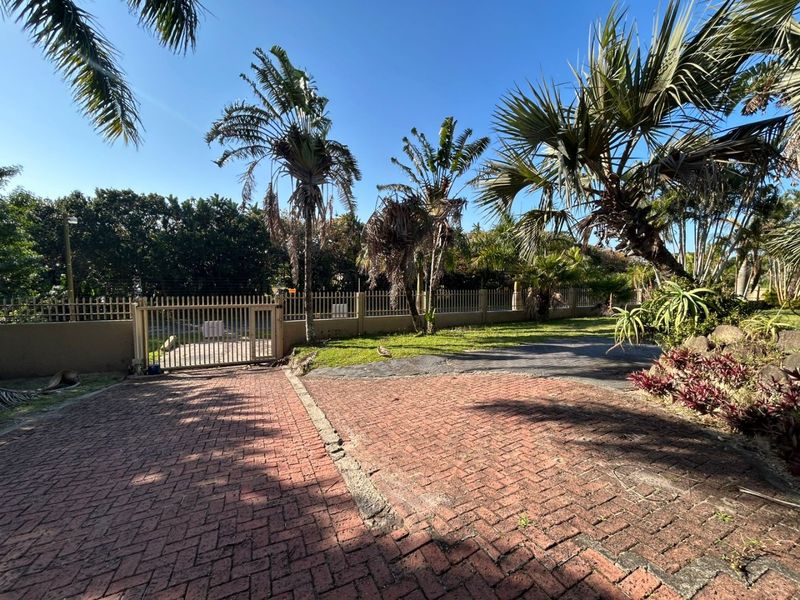 A front exterior view featuring a paved driveway, metal security gate, and established garden with perimeter fencing.