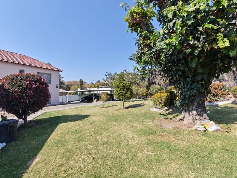 A well-maintained garden area featuring a lush lawn, mature trees, and a paved driveway leading to a carport.