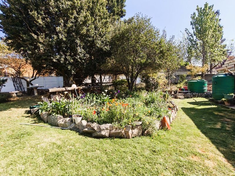 A well-established garden featuring a stone-edged flower bed, mature trees, and a green water storage tank set within a private backyard area.