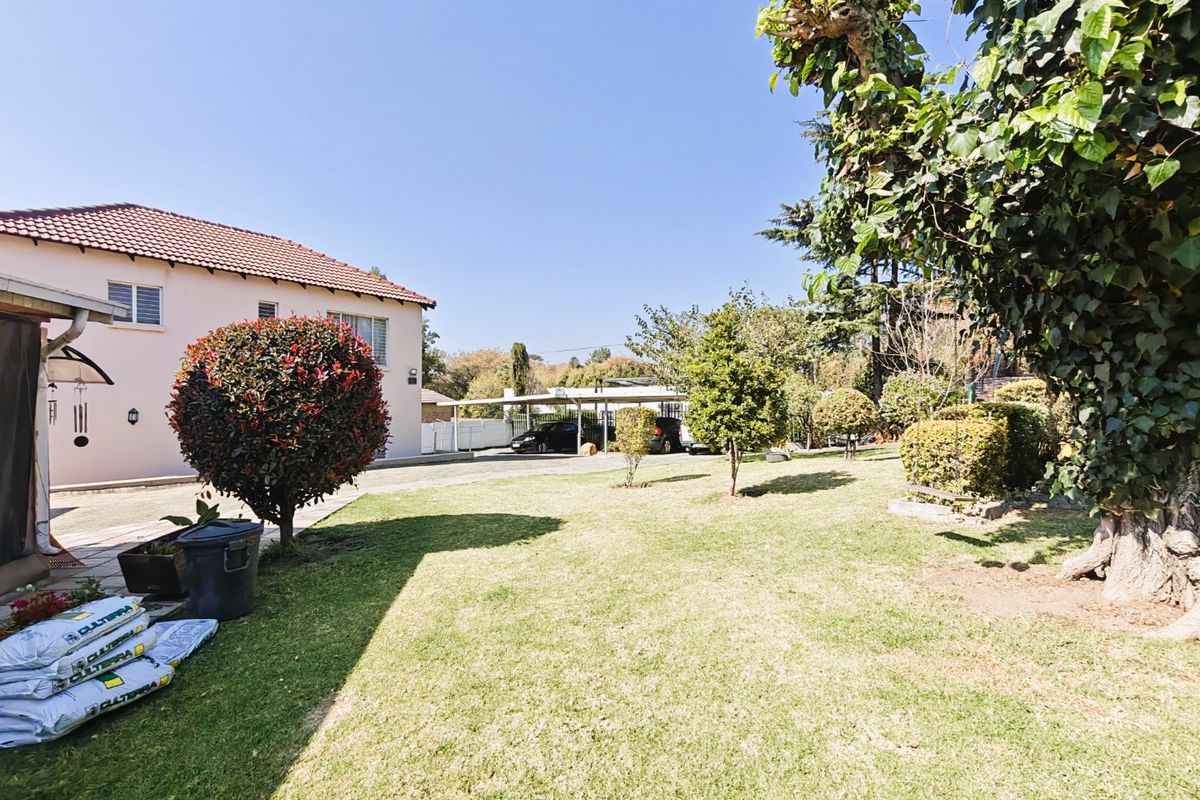 A spacious rear garden featuring a well-maintained lawn, mature trees, and a detached carport structure visible in the background.