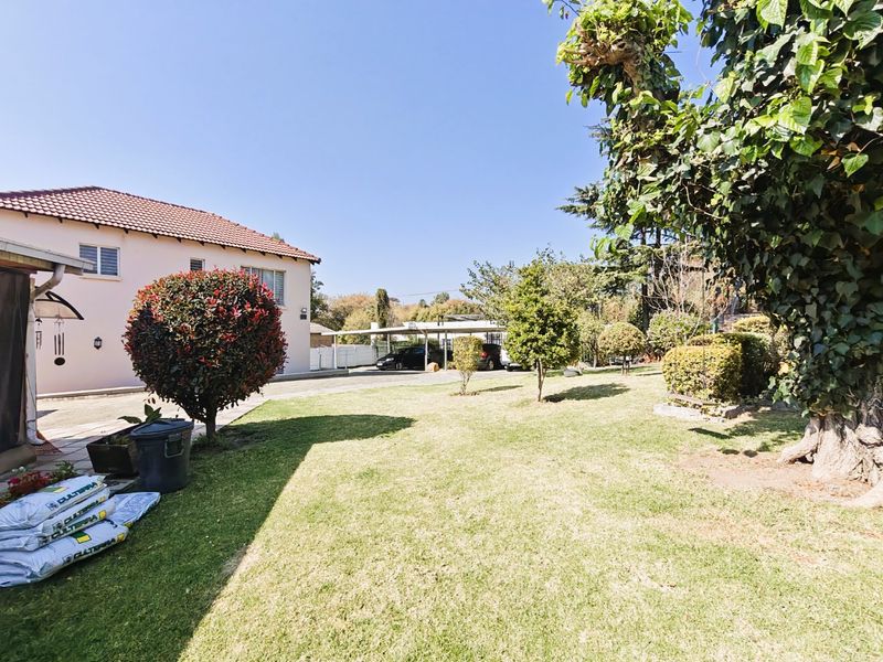 A spacious rear garden featuring a well-maintained lawn, mature trees, and a detached carport structure visible in the background.
