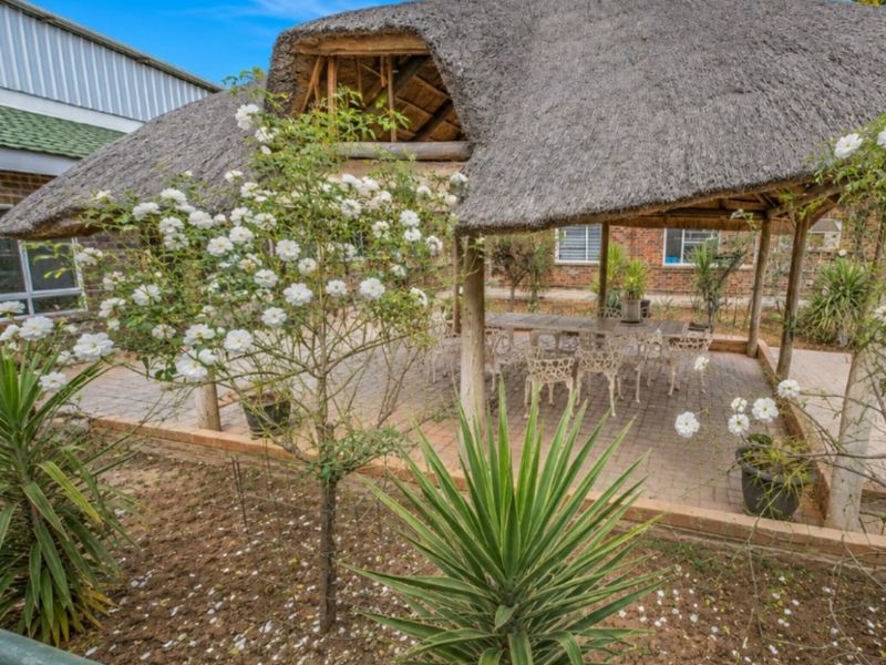 A covered outdoor patio area featuring a traditional thatched roof structure and brick paving, set within a landscaped garden environment.