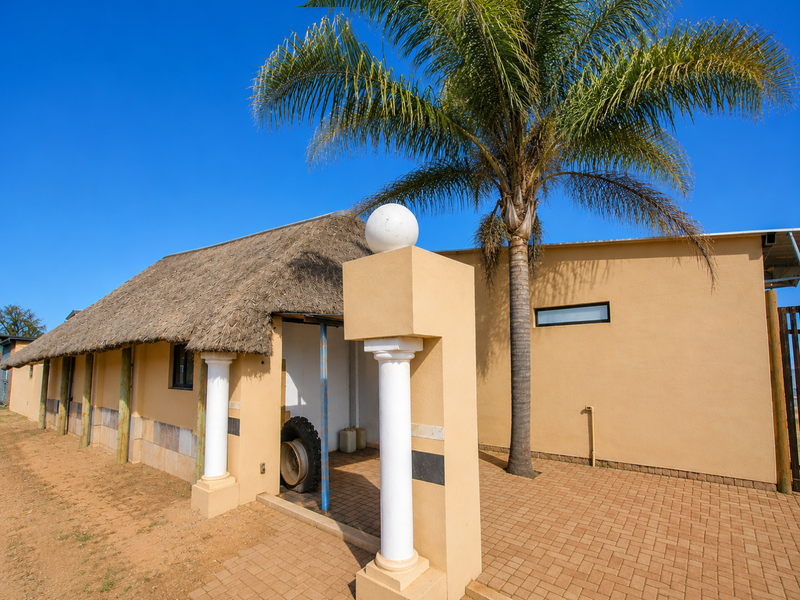 Front exterior view of a commercial building featuring a traditional thatch roof, decorative columns, and paved entrance area with established landscaping.