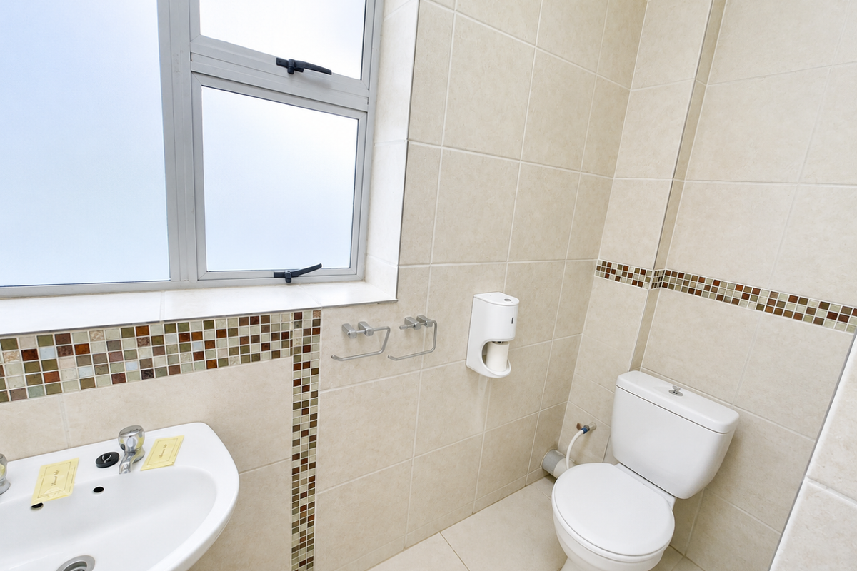 A clean, functional bathroom featuring a pedestal washbasin, toilet, neutral wall tiles with a mosaic accent border, and a frosted glass window.