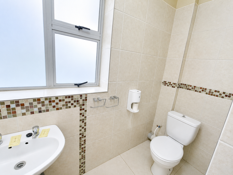 A clean, functional bathroom featuring a pedestal washbasin, toilet, neutral wall tiles with a mosaic accent border, and a frosted glass window.