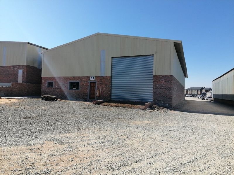 Industrial warehouse unit featuring a large roller shutter door, brick and metal cladding exterior, and a spacious gravel yard area.