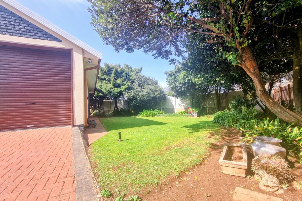 A well-maintained rear garden featuring a lush lawn, mature trees, and a paved driveway leading to a garage with a roller door.