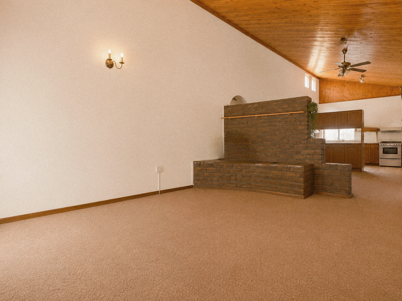 Spacious open-plan living and kitchen area featuring a vaulted timber ceiling, brick feature wall, and neutral carpeting throughout.