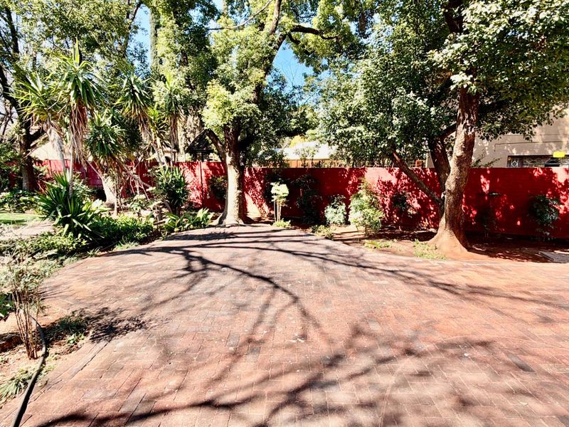 A spacious outdoor garden area featuring a large brick-paved patio, mature trees, and a solid boundary wall.