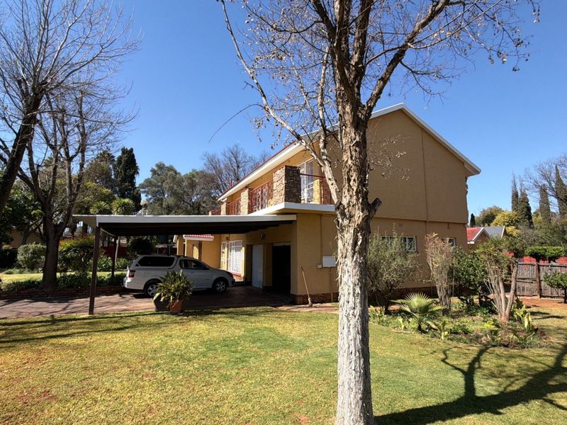 Side profile of a two-story house featuring a carport, paved driveway, and an upper-level balcony with stone detailing, set within a landscaped garden.