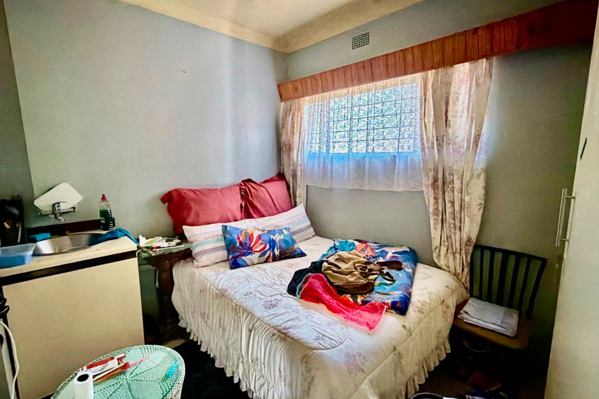 A compact bedroom featuring a built-in sink, security bars on the window, and a ventilation grille, requiring cosmetic updates to improve its overall condition.