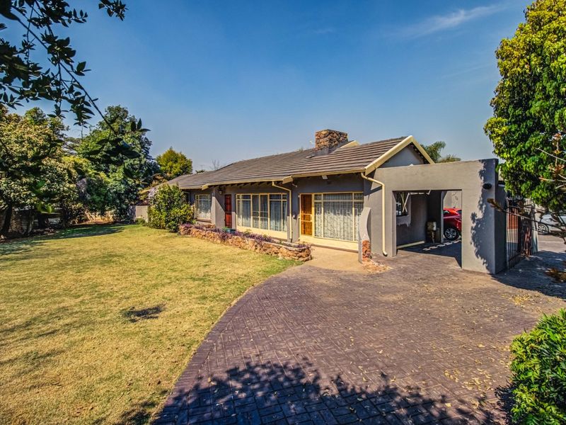 Front view of a single-story house featuring a paved driveway, attached carport, stone retaining wall, and a spacious front garden.