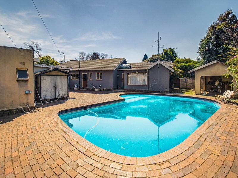 Rear exterior view featuring a swimming pool, brick-paved patio area, and adjacent outbuildings under a clear sky.
