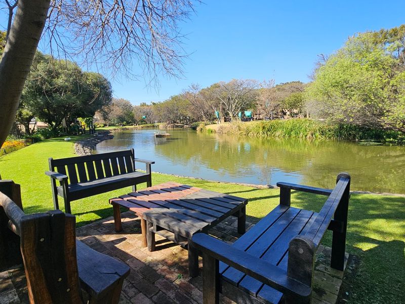 A scenic view of a tranquil pond surrounded by a well-maintained garden and mature trees, featuring a paved outdoor seating area.