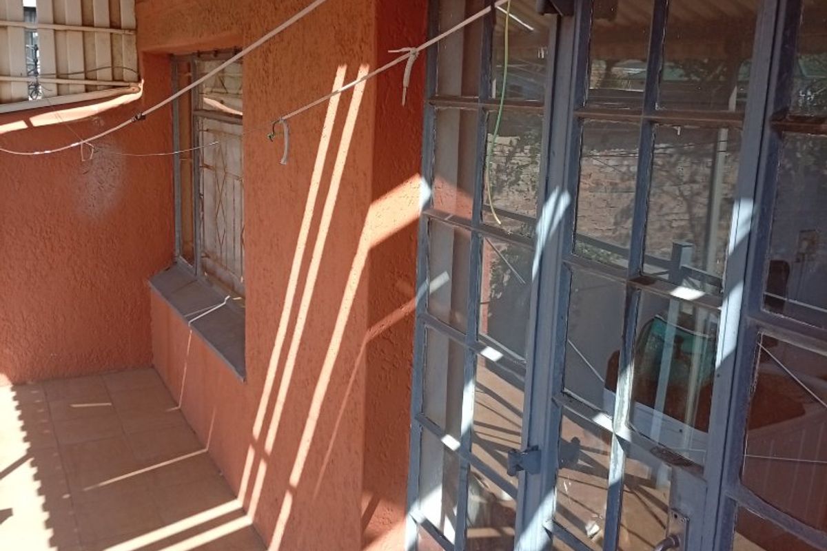A covered patio area featuring tiled flooring, steel-framed glass doors, and textured exterior walls, offering a functional outdoor space in need of maintenance.