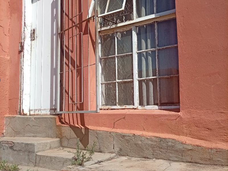 Exterior view of a building entrance featuring concrete steps, a security gate, and a steel-framed window with visible signs of wear and structural cracking.
