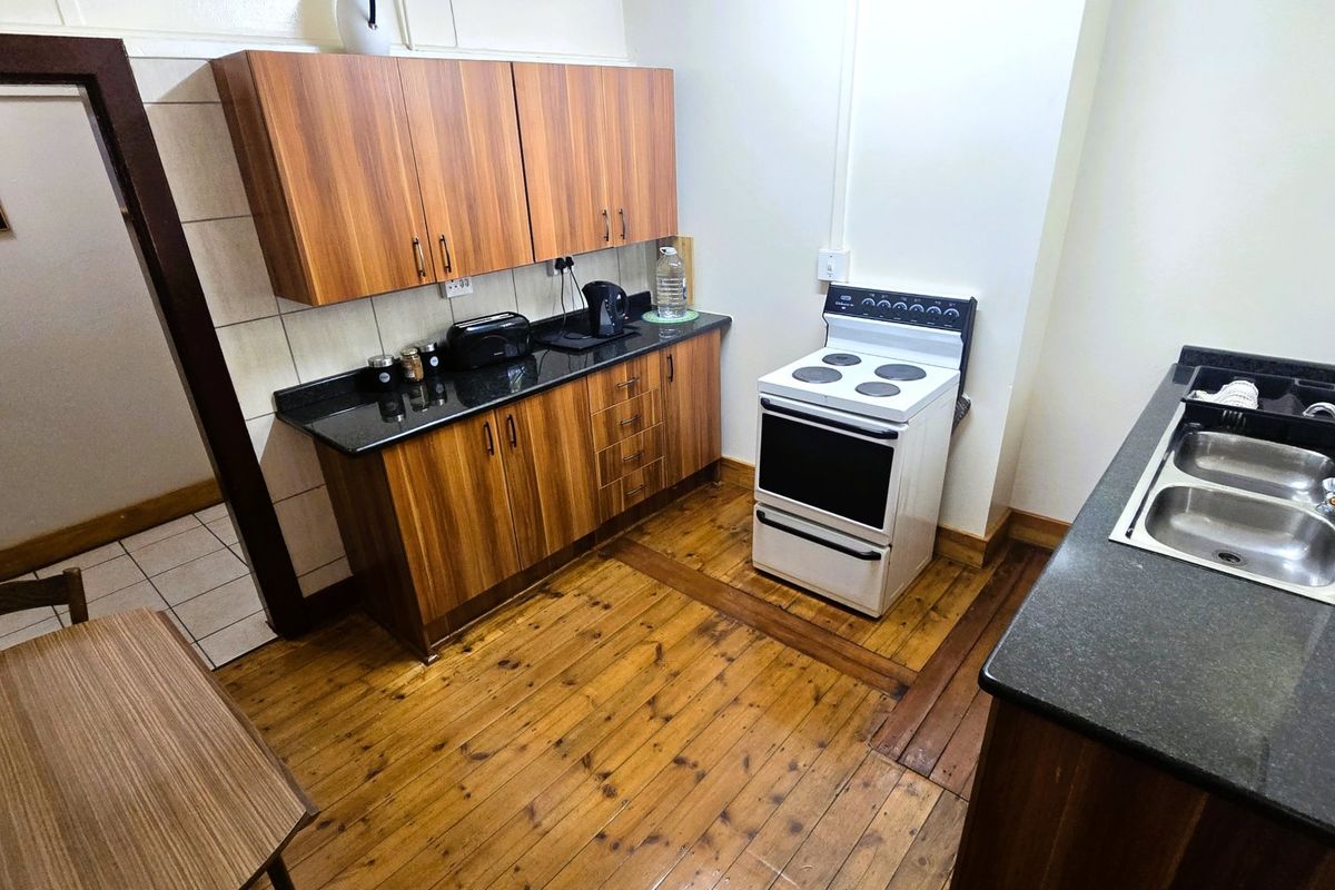 A functional kitchen featuring wood cabinetry, granite countertops, a freestanding electric stove, and a double stainless steel sink with timber flooring.