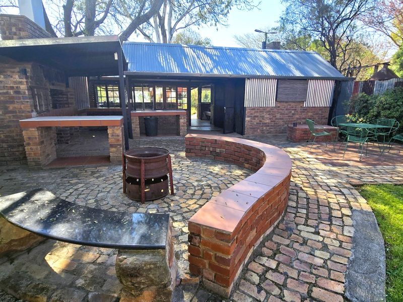 Outdoor entertainment area featuring a brick built-in braai, curved seating wall, and cobblestone paving under a corrugated metal roof structure.